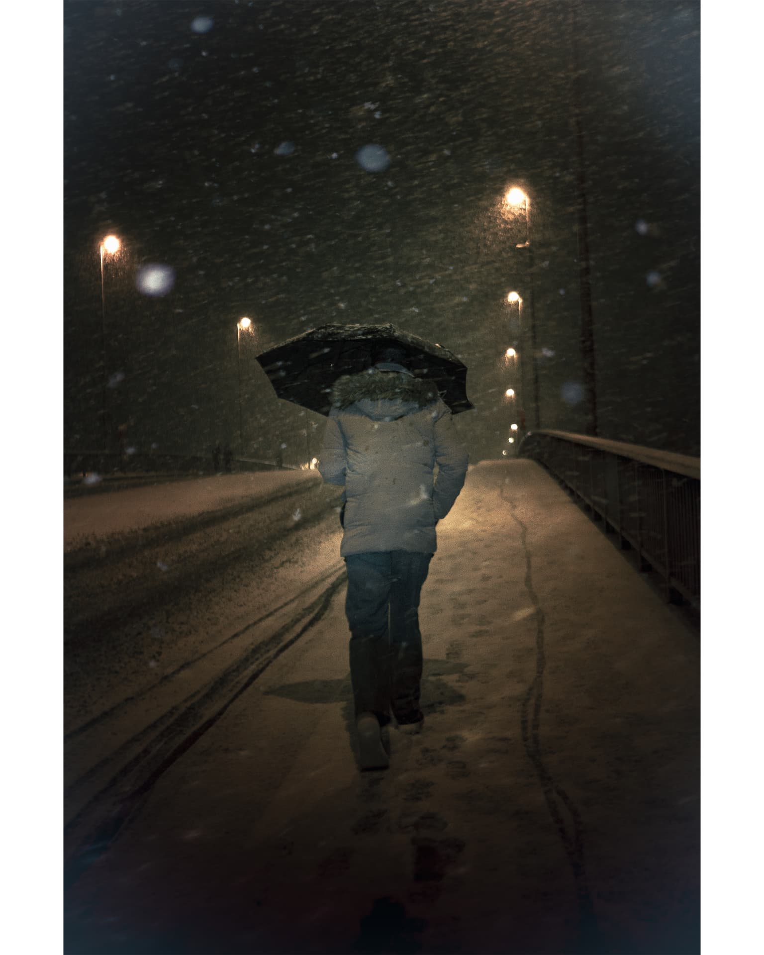 A person walking away under a dark umbrella along a snowy bridge at night